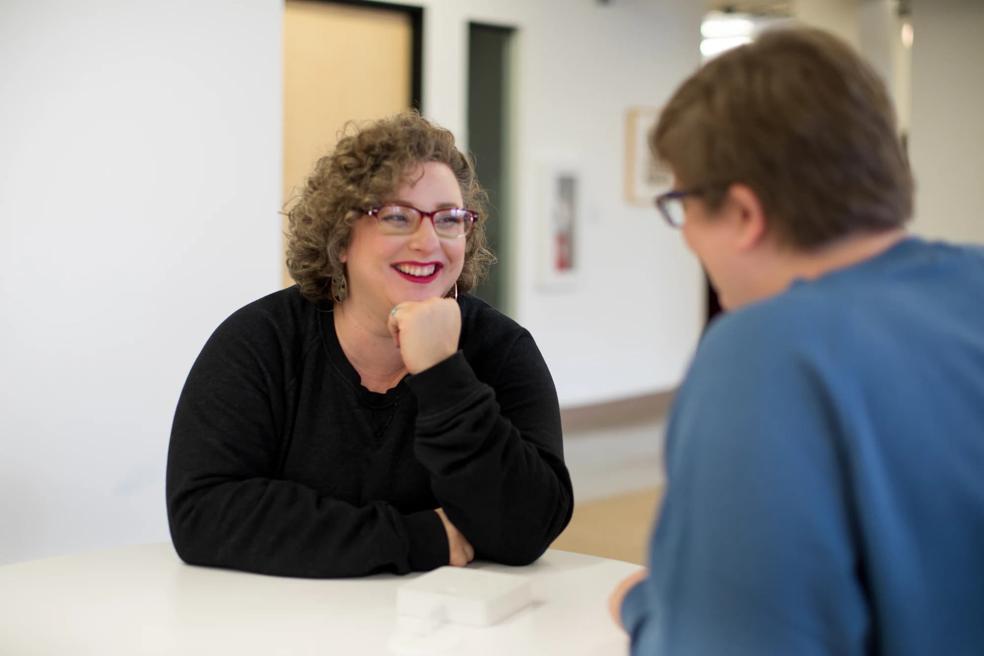Two people talking across a table in a bright room, one of them laughing.