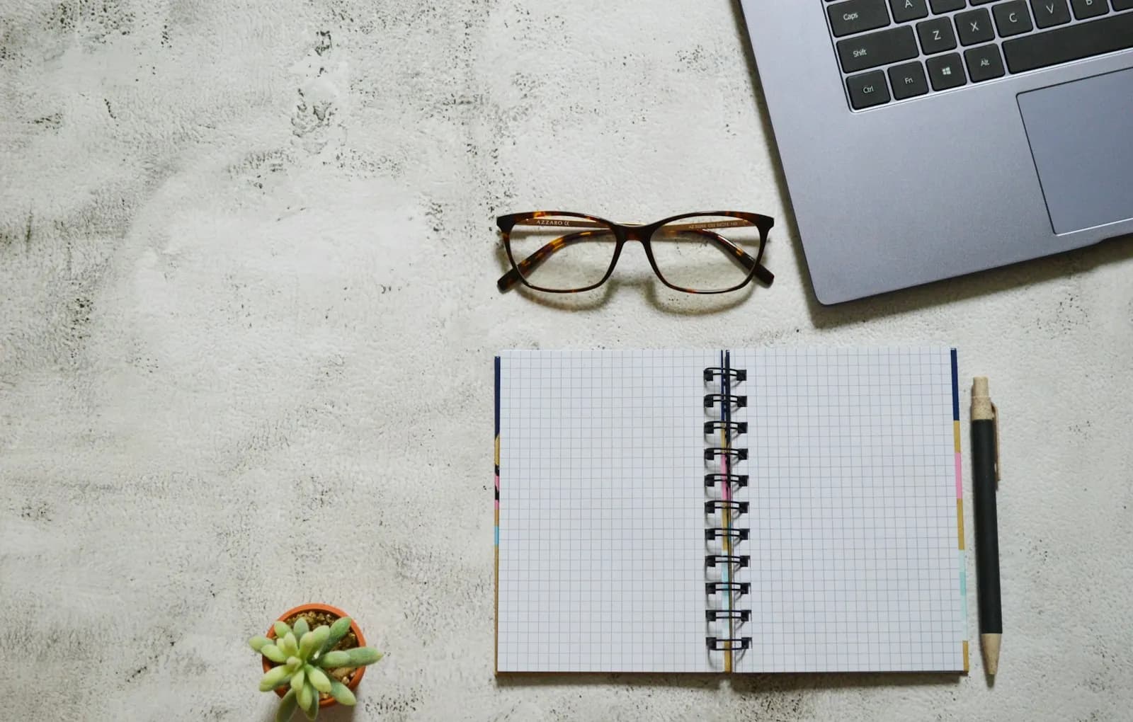 A notebook, glasses and a laptop on a desk, ready for writing interface copy.